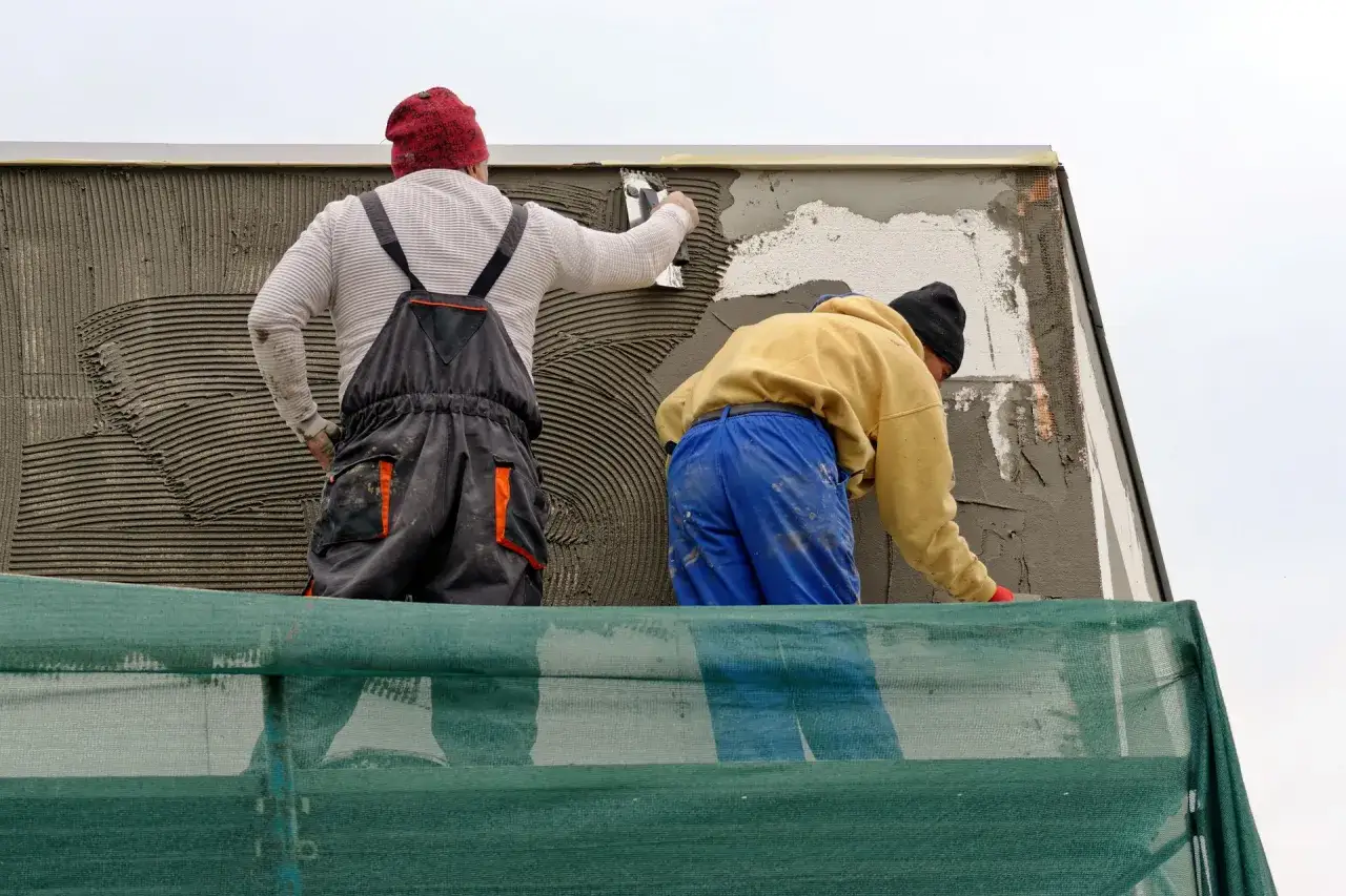 Lavoratori impegnati nella rasatura di un muro esterno, applicano l'intonaco con spatole su un'impalcatura.