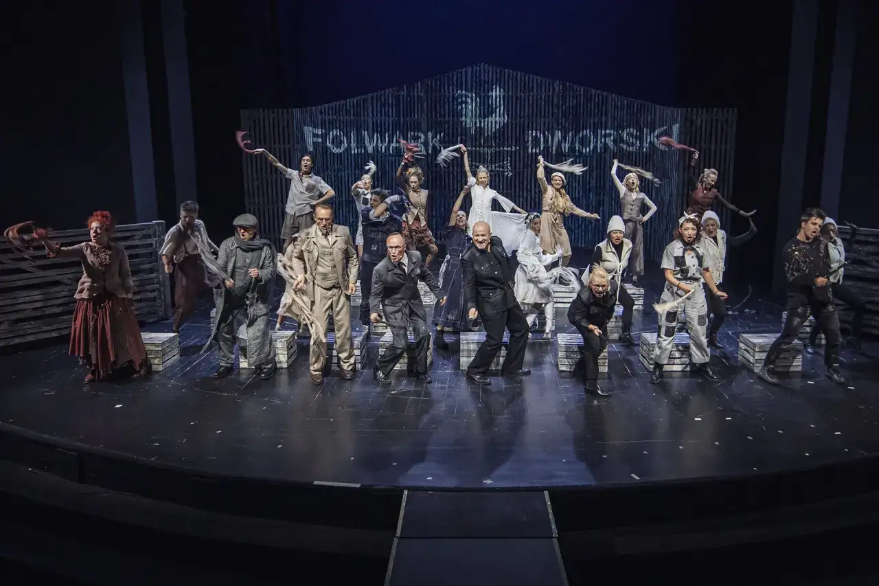 Aktorzy w strojach z epoki odgrywają scenę w teatrze. W tle widnieje napis 