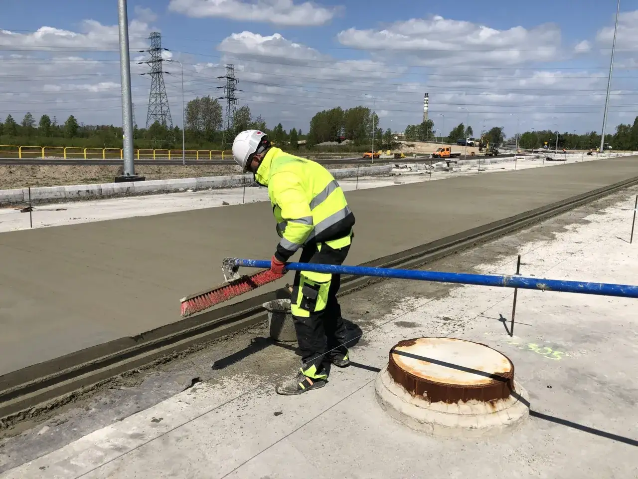 Pracownik w kamizelce odblaskowej wygładza świeży, mokry beton. W tle widać linie energetyczne i maszyny budowlane.