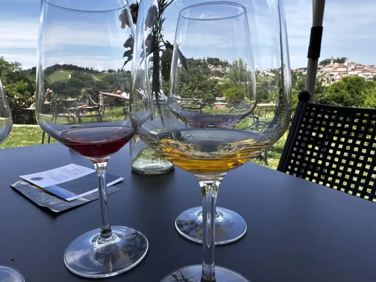 Calice di vino rosso e bianco, con un paesaggio collinare sullo sfondo. Sembra che manchi solo un quinto per completare la degustazione.