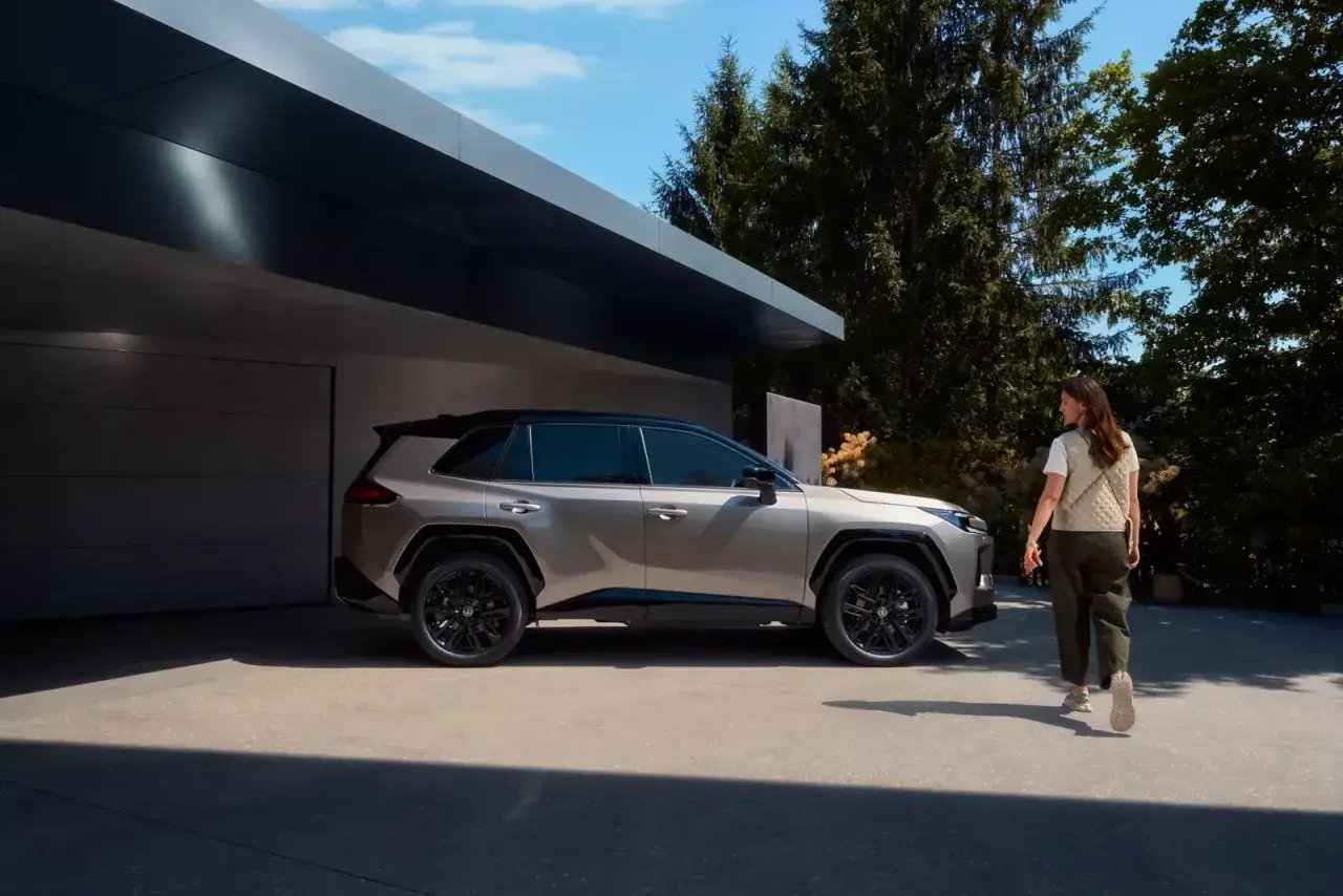 Elegancka kobieta zbliża się do nowego **Toyota RAV4 2025** zaparkowanego przed nowoczesnym domem.