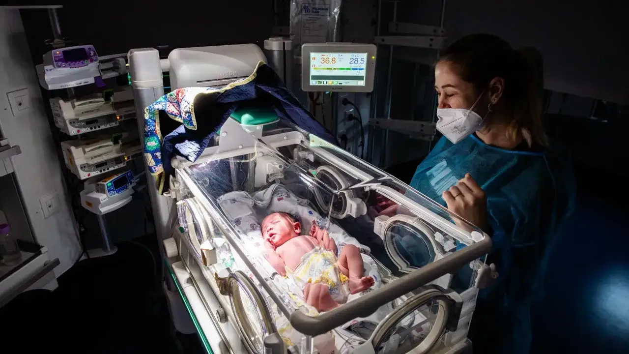 Un neonato dorme in una culla termica nella terapia intensiva neonatale, mentre un'infermiera lo osserva con cura.