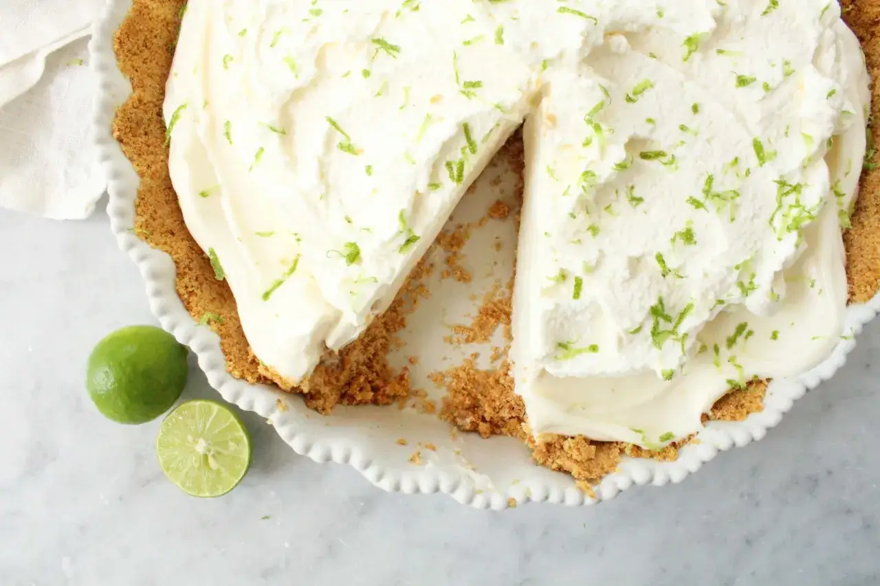 En kr&auml;mig key lime pie med en krispig botten, toppad med vispad gr&auml;dde och rivet limeskal. En hel och en halv lime ligger bredvid.
