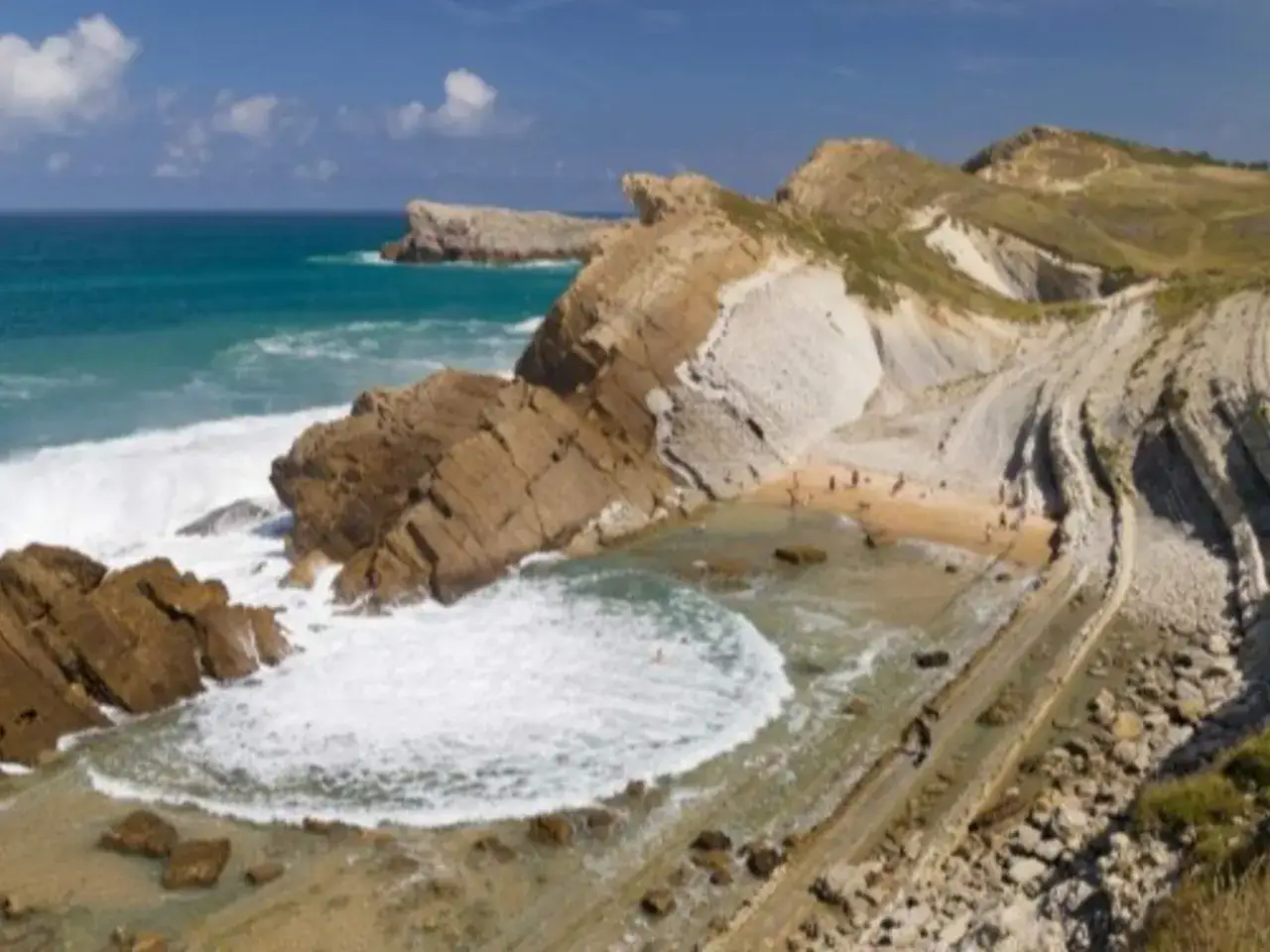 Olas rompen en una cala rocosa, una **playa de piezas** geológicas únicas con capas de roca expuestas y gente disfrutando del sol y el mar.