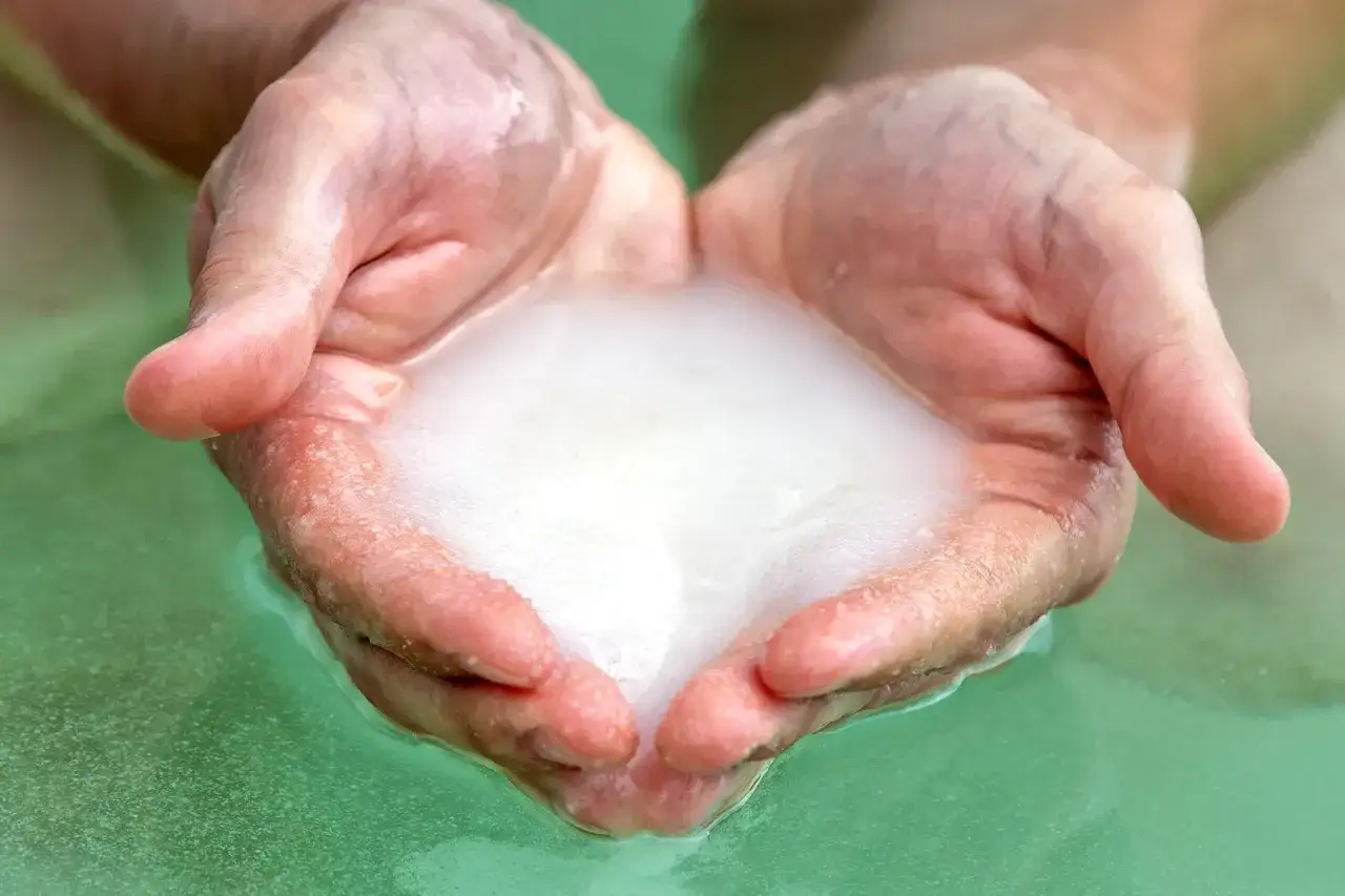 Mani che raccolgono schiuma bianca in acqua verde, evocando i benefici dei sali del Mar Morto per la pelle.
