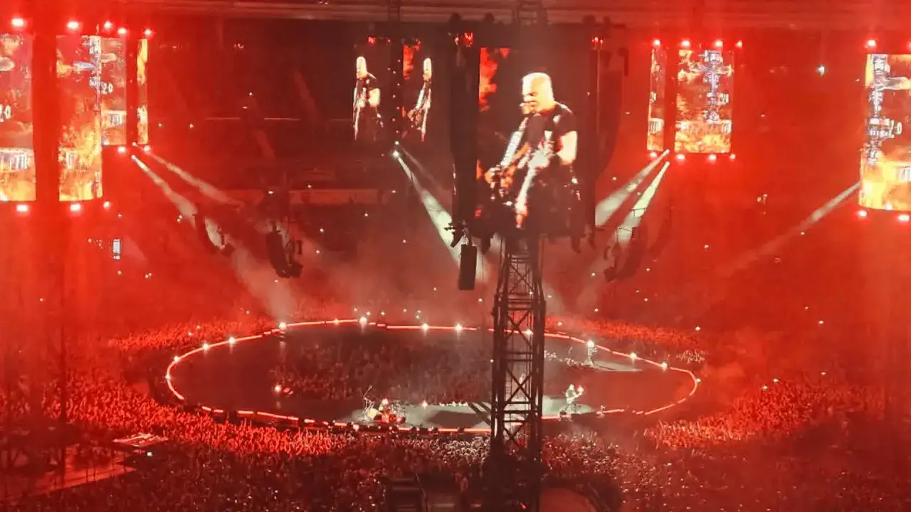 Koncert rockowy w czerwonym świetle. Na ekranach widać muzyka, a tłum na widowni uśmiecha się.