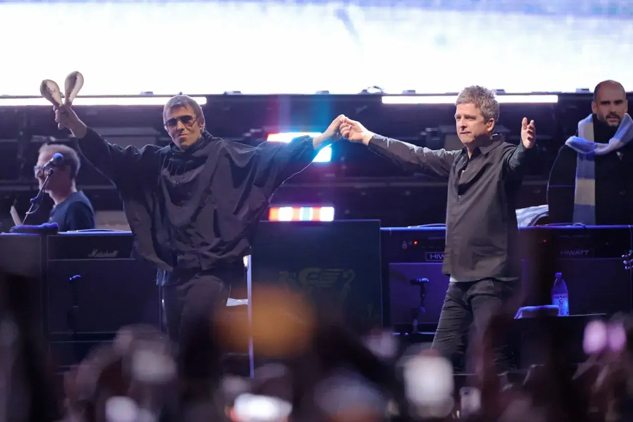 Les frères Gallagher, membres de Oasis, se tiennent la main sur scène. Liam agite des maracas, Noel tend les bras.