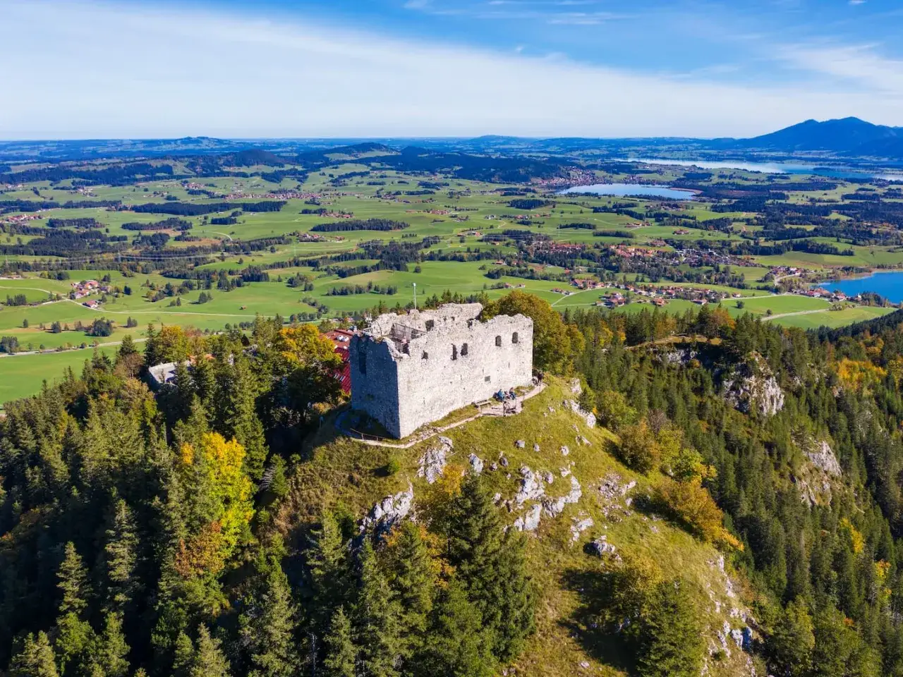 Ruinen der Burg Falkenstein bei Pfronten thronen auf einem H&uuml;gel, umgeben von W&auml;ldern und mit Blick auf eine malerische Alpenlandschaft mit Seen.