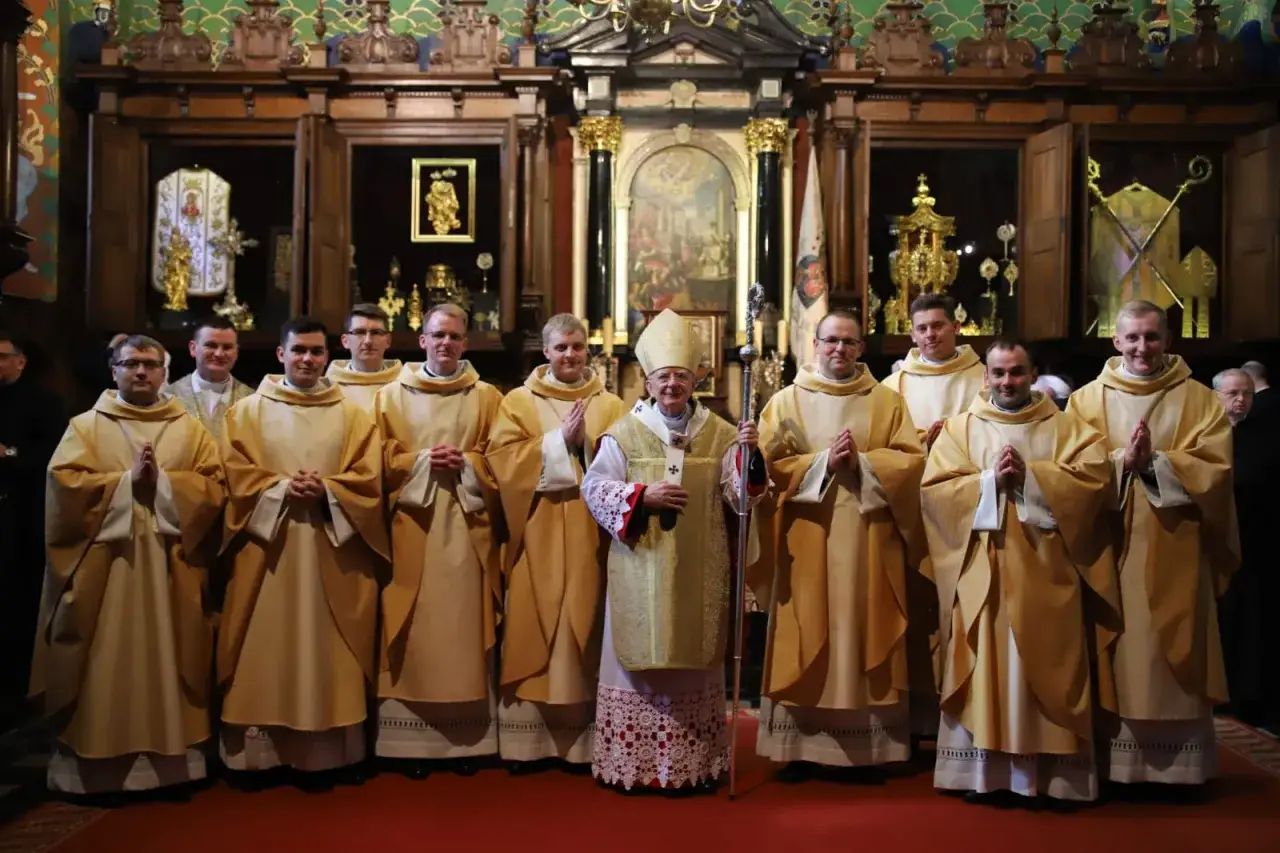 Biskup w złotej szacie stoi pośród grupy prezbiterów w kościele.
