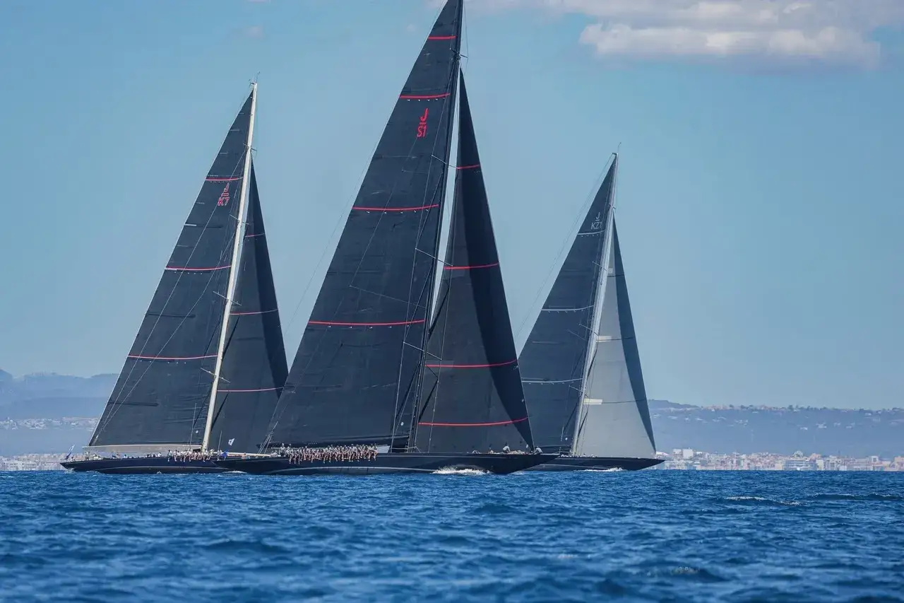 Tre imbarcazioni J class sfrecciano sull'acqua blu, con vele scure e rosse che catturano il vento.