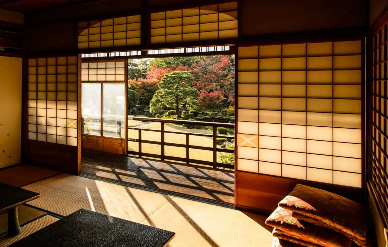 Intérieur d'une maison japonaise avec des cloisons shoji. Le soleil filtre à travers, créant des ombres sur les tatamis. Dehors, un jardin d'automne aux couleurs vives.
