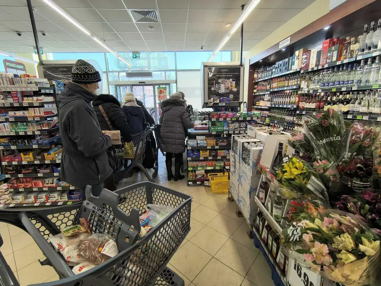 Klienci w kolejce do kasy w sklepie Dino. W wózku zakupy, na półkach słodycze i alkohol.