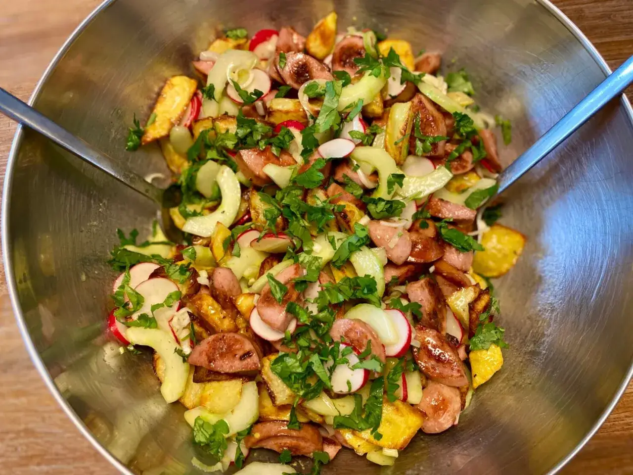 Ein herzhafter, **crunchy Kartoffelsalat** mit Wurstscheiben, Radieschen und Gurke, garniert mit frischer Petersilie in einer Edelstahlsch&uuml;ssel.