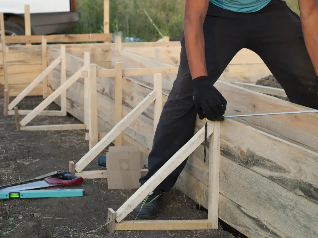 Un ouvrier mesure le coffrage en bois pour le futur petit coffrage béton. Des outils sont posés au sol.