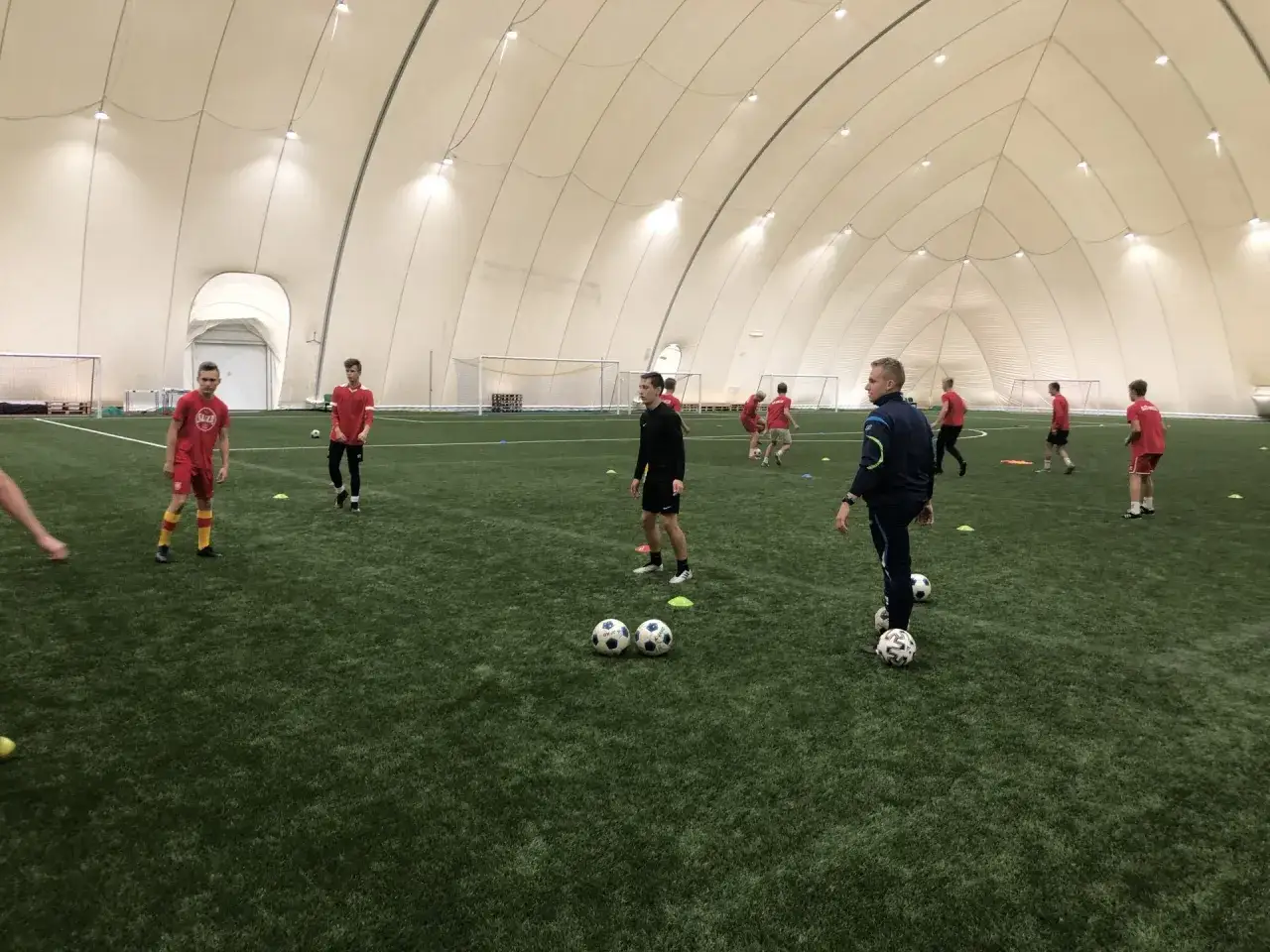 Trening piłkarski w hali. Młodzi zawodnicy w czerwonych strojach ćwiczą pod okiem trenera, spełniając **uefa c wymagania**.