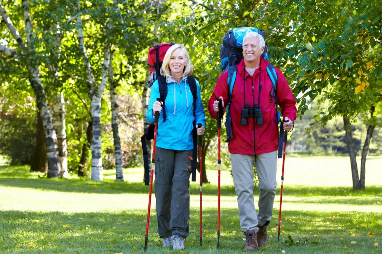 Uśmiechnięta para starszych ludzi z plecakami i kijami do **nordic walking** spaceruje po zielonej trawie wśr&oacute;d drzew.