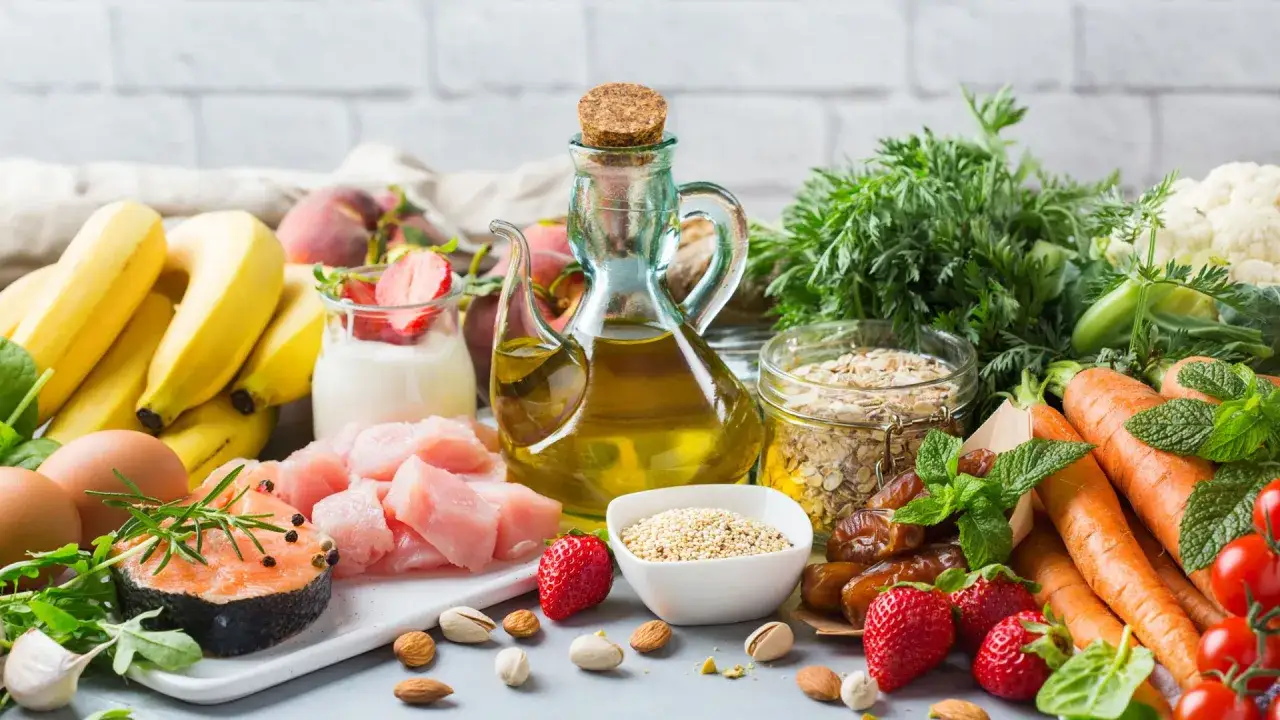 Variedad de alimentos frescos: salmón, pollo, huevos, frutas (plátanos, fresas, melocotones), verduras (zanahorias, coliflor, espinacas), frutos secos y aceite de oliva, todos ricos en vitaminas para piel radiante.