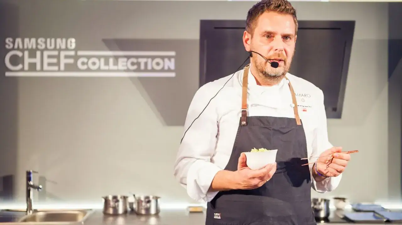 Szef kuchni Amaro, w skromnym fartuchu, prezentuje składniki na tle logo Samsung Chef Collection.