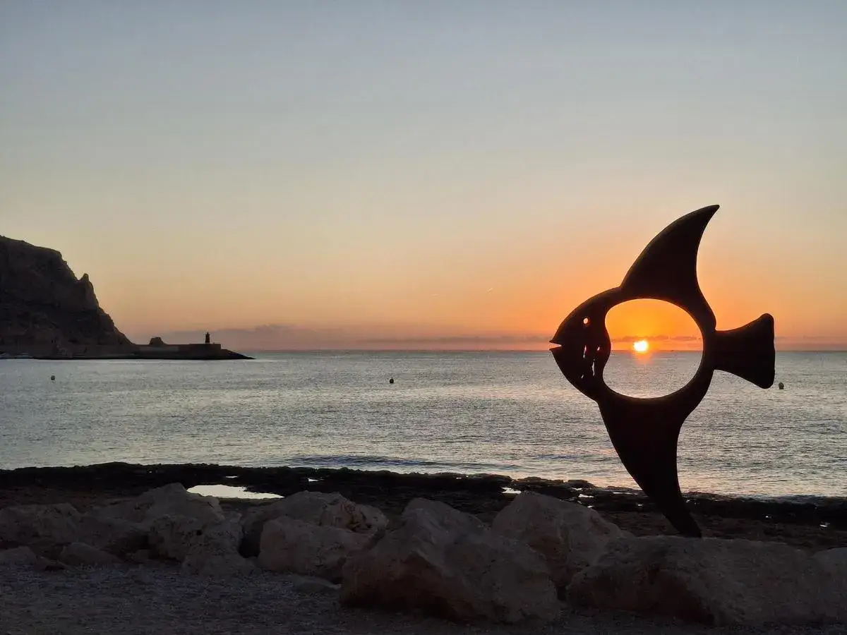 Escultura de pez silueteada contra un amanecer en el mar. Parece que está a punto de soñar con peces fuera del agua, contemplando el sol.