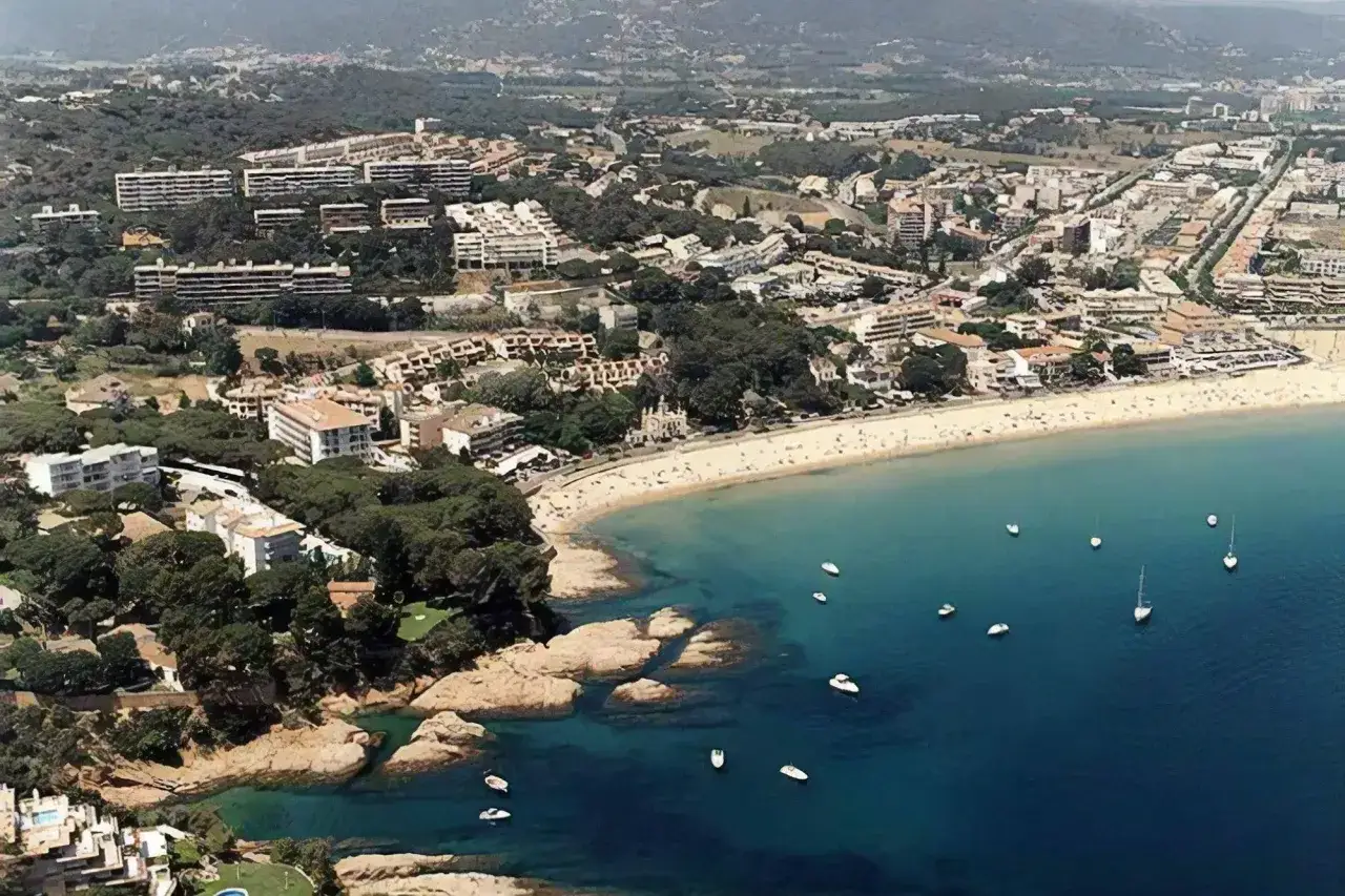 Vista aérea de la platja Sant Pol en Sant Feliu de Guíxols, con edificios, árboles y barcos en el mar azul.