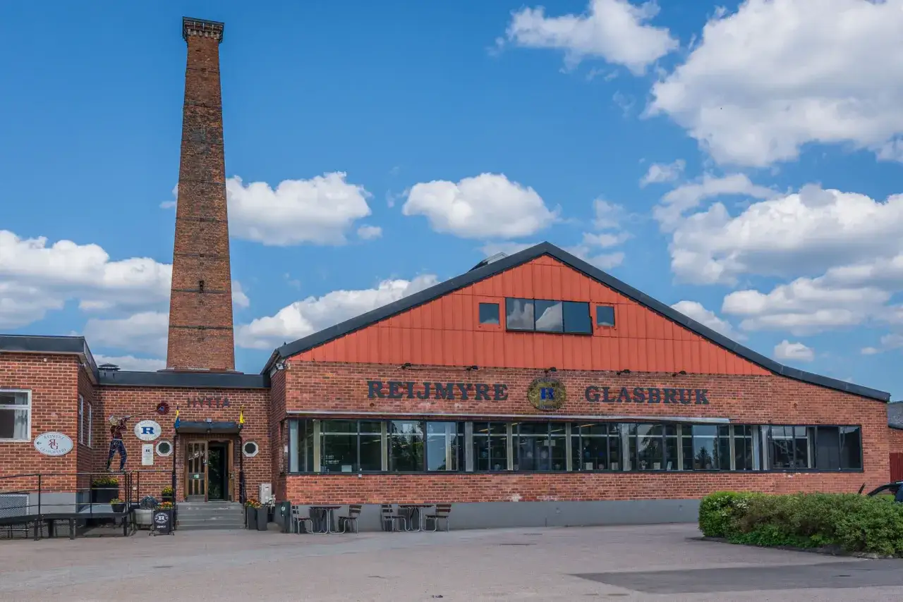 Reijmyre Glasbruk, en del av Rejmyre hantverksby, med sin höga tegelpipa under en blå himmel med moln.