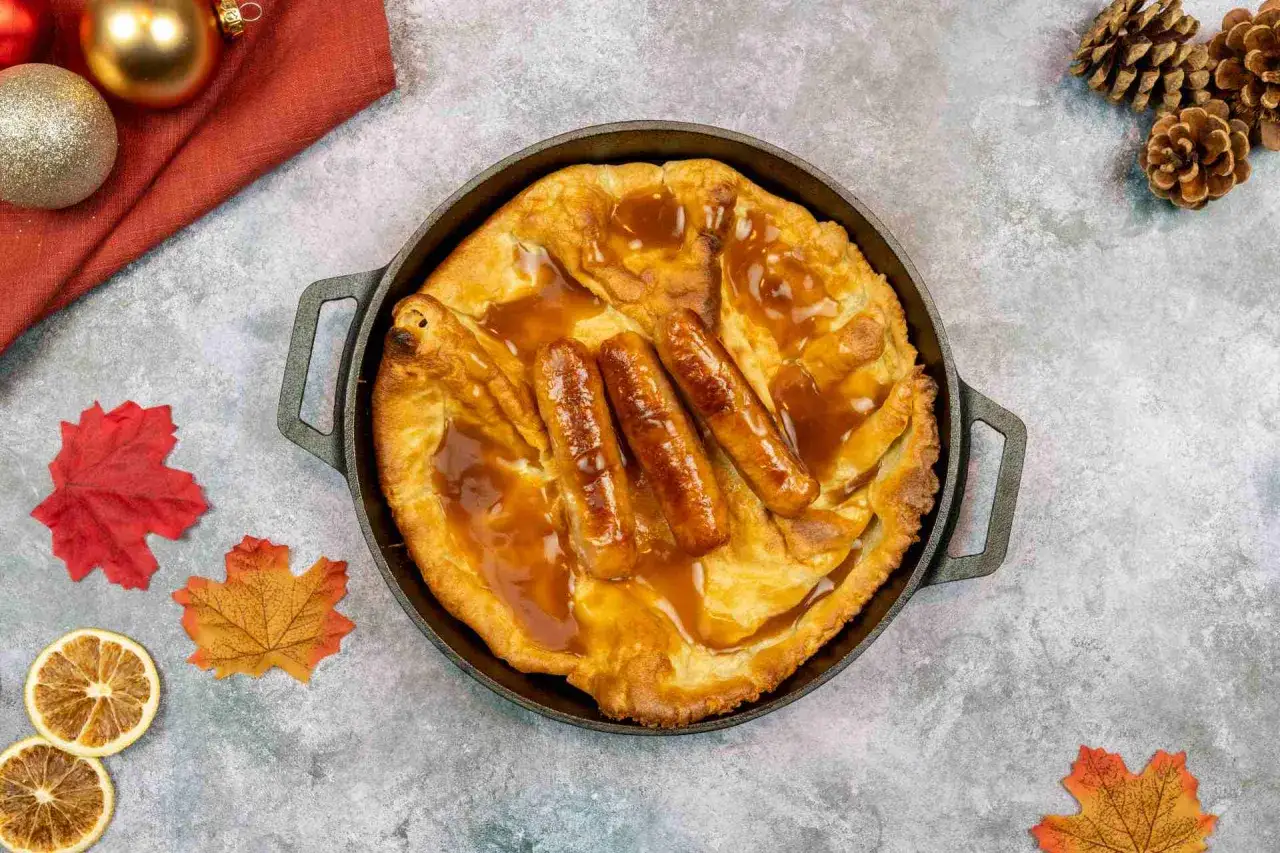 Herzhaftes Yorkshire Pudding Rezept mit Würstchen und brauner Soße in einer Gusseisenpfanne, dekoriert mit Herbstblättern und Tannenzapfen.