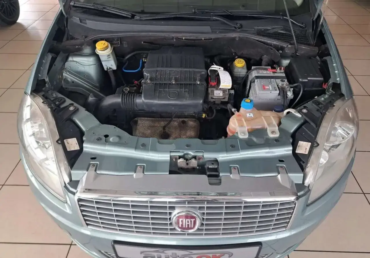 Silnik w jasnoniebieskim Fiat Linea. Widoczny akumulator, zbiorniczki płynów i elementy układu dolotowego.
