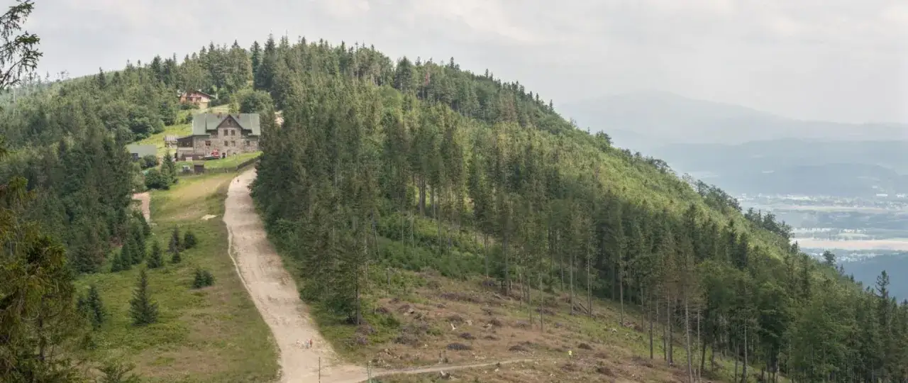 Na zboczu góry, porośniętej lasem, widać kamienny budynek schroniska. Droga prowadzi w górę, a w oddali majaczą kolejne, mgliste szczyty. To wielki stożek górski.