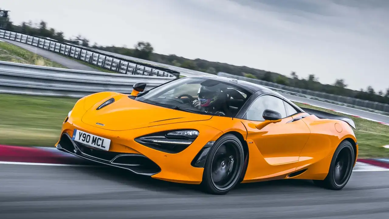 Pomarańczowy McLaren 720S z kierowcą w kasku pędzi po torze wyścigowym, zostawiając za sobą smugę prędkości.