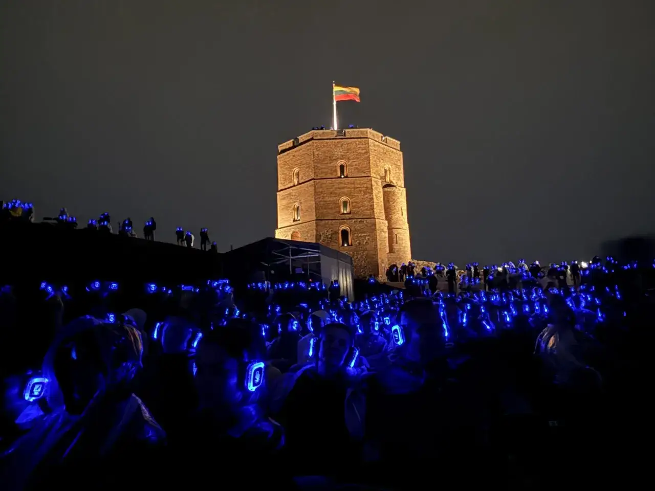Nocny koncert w słuchawkach przed wieżą w Wilnie. Tłum ludzi z niebieskimi słuchawkami tworzy świetlisty dywan. Stare miasto Wilno nocą.