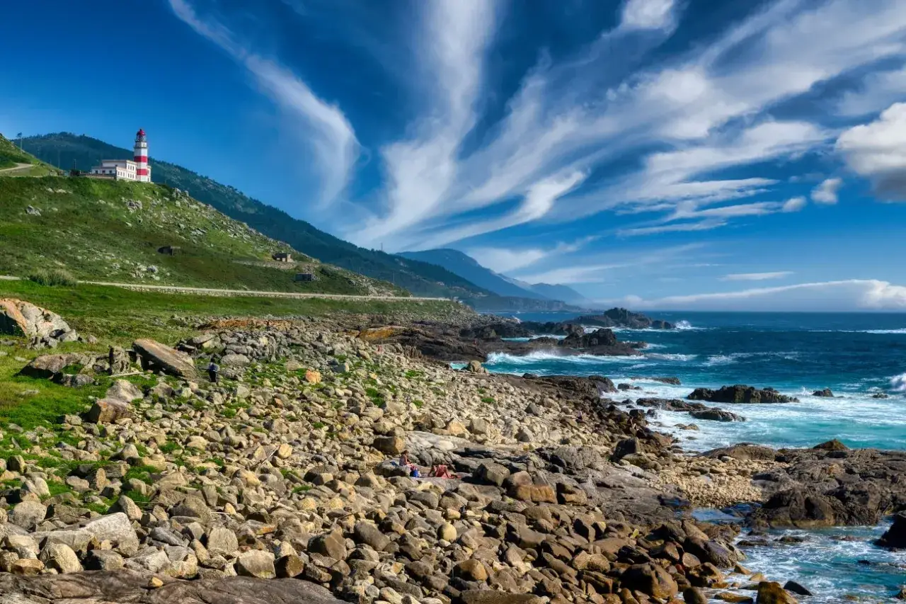 El faro de Baiona se alza en una colina verde junto a un mar azul y rocoso. Nubes blancas y finas surcan el cielo.