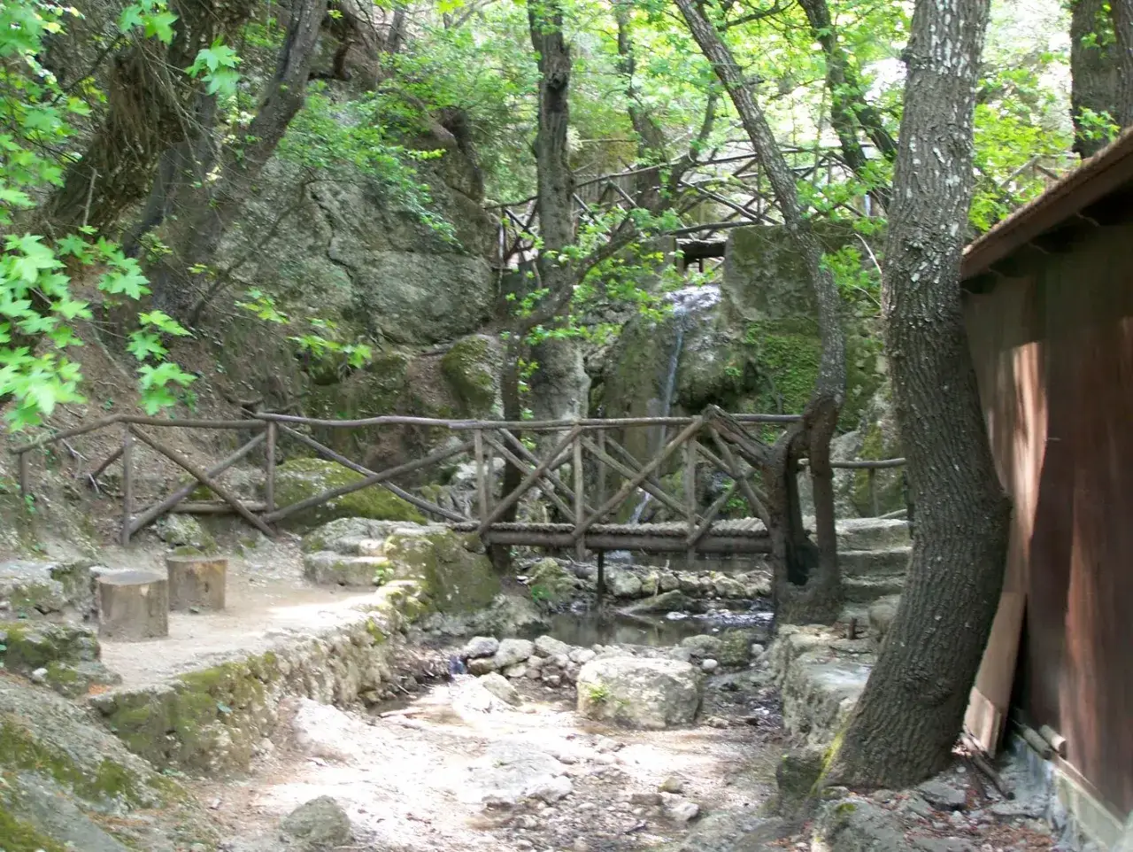 Holzbr&uuml;cke im Schmetterlingstal auf Rhodos, &uuml;ber einem Bachlauf, umgeben von &uuml;ppiger Vegetation und Felsen.