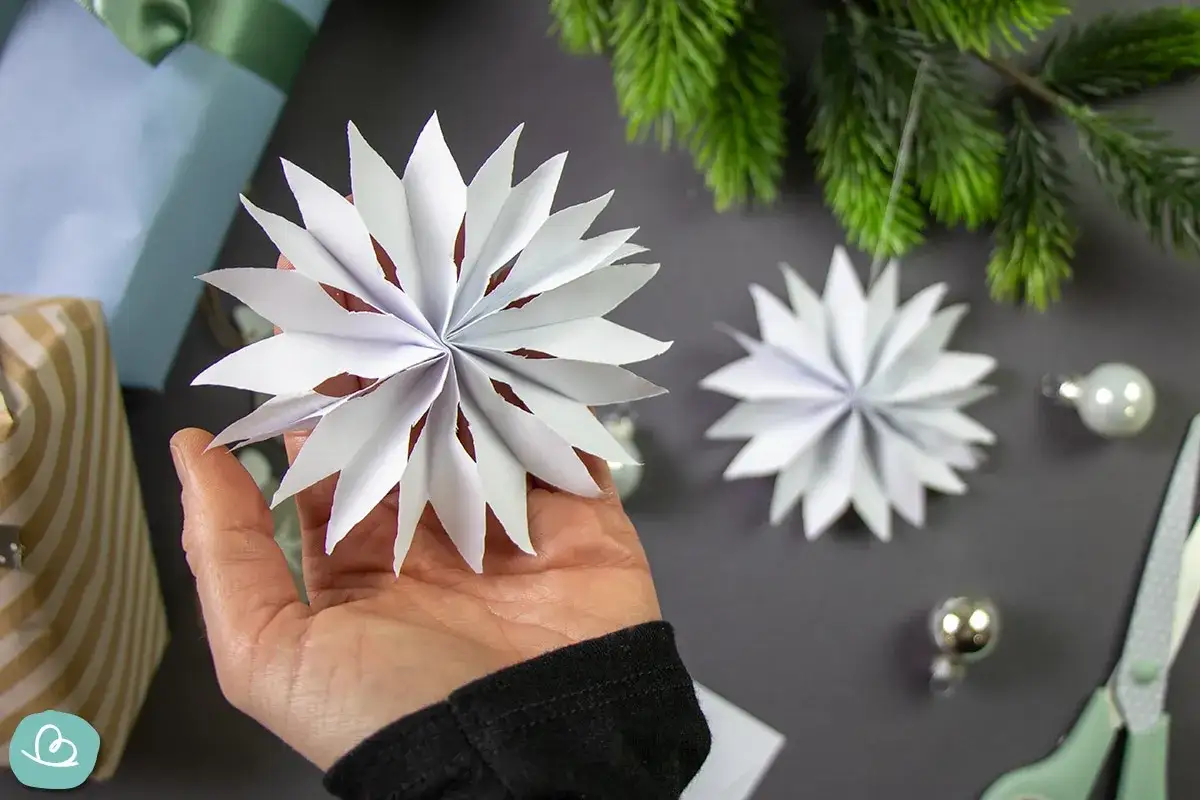 Hand hält selbstgemachten DIY Christbaumschmuck aus Papier. Daneben liegen Geschenke, Tannenzweige und Kugeln.