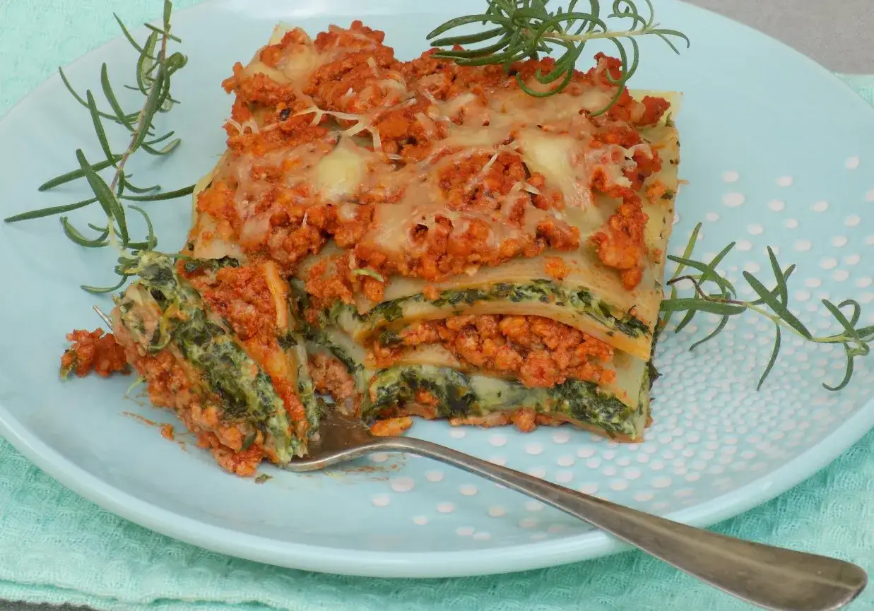 Prosty przepis na lasagne z mięsem mielonym i szpinakiem, podana na błękitnym talerzu, ozdobiona gałązkami rozmarynu.