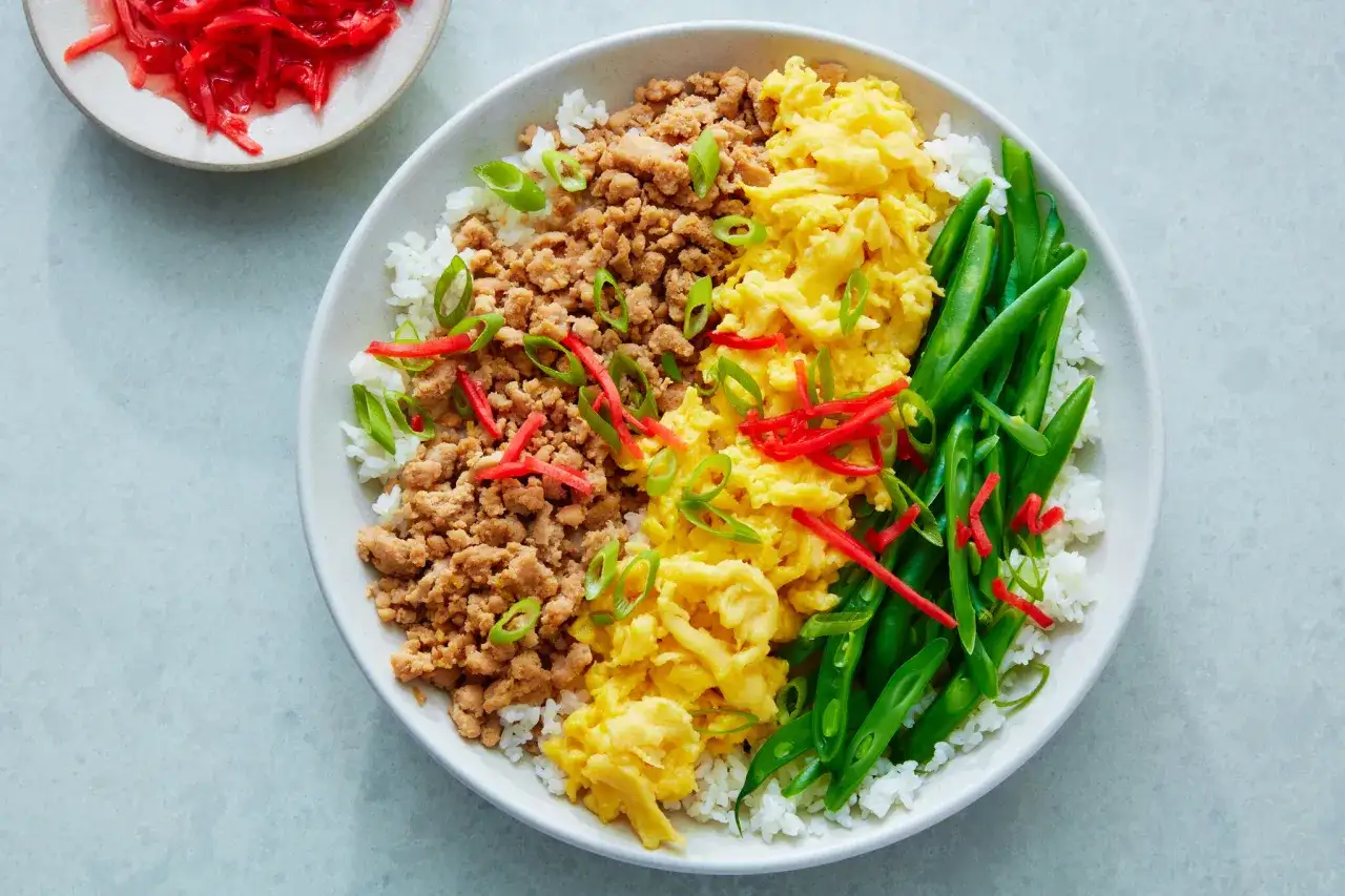 A colorful sanshoku don with rice, seasoned ground meat, scrambled eggs, and green beans, garnished with scallions and red ginger.