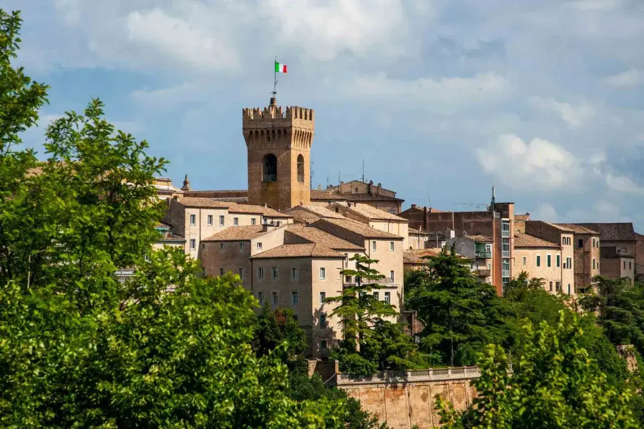 La città di Leopardi, con la sua torre medievale e le case in pietra, si staglia tra il verde degli alberi sotto un cielo azzurro e nuvoloso.