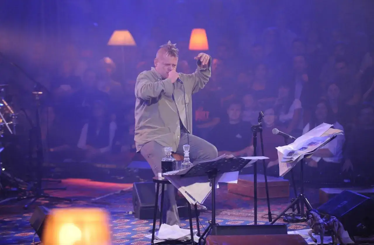 Kazik Staszewski podczas MTV Unplugged Polska, siedząc na stołku, śpiewa do mikrofonu. W tle widać publiczność i lampy.