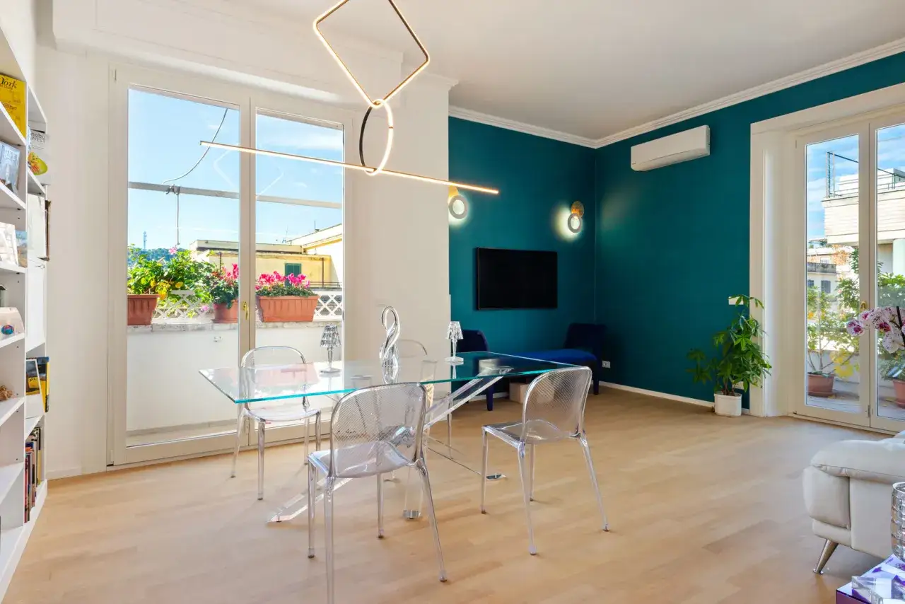 Un soggiorno moderno con pareti color blu pavone, un tavolo in vetro e sedie trasparenti.