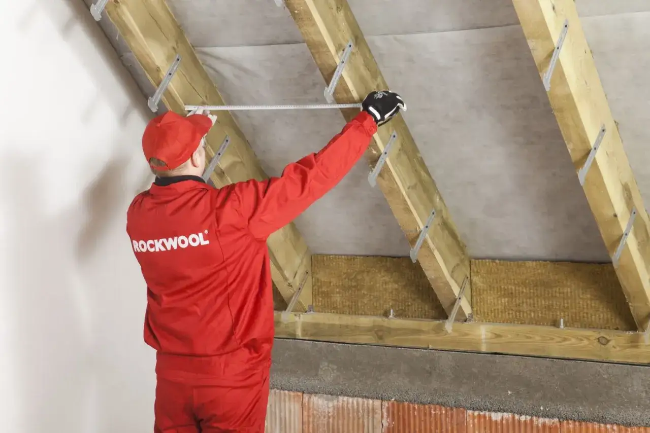 Pracownik w czerwonym kombinezonie Rockwool mierzy przestrzeń między krokwiami, by dobrać odpowiednią grubość wełny do izolacji poddasza.