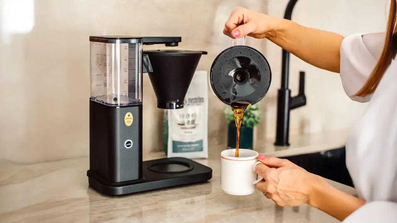 En kaffebryggare i k&ouml;ket. En kvinna h&auml;ller upp nybryggt kaffe i en mugg. Enkel guide f&ouml;r hur brygger man kaffe.