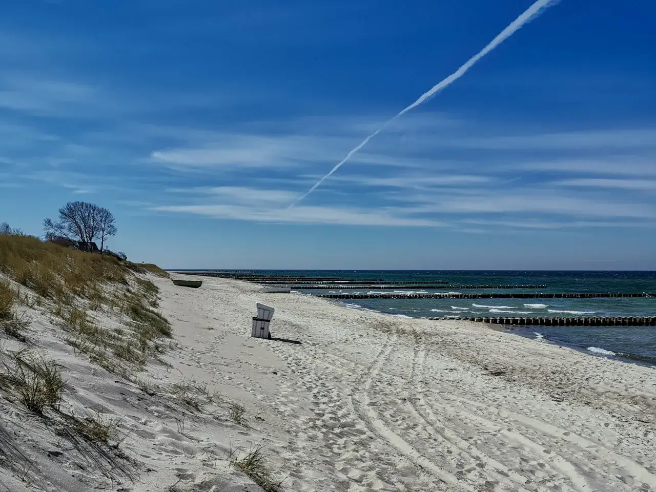 Ein einsamer Strandkorb am Ahrenshoop Strand, mit Dünen, dem Meer und einer Flugzeugspur am blauen Himmel.