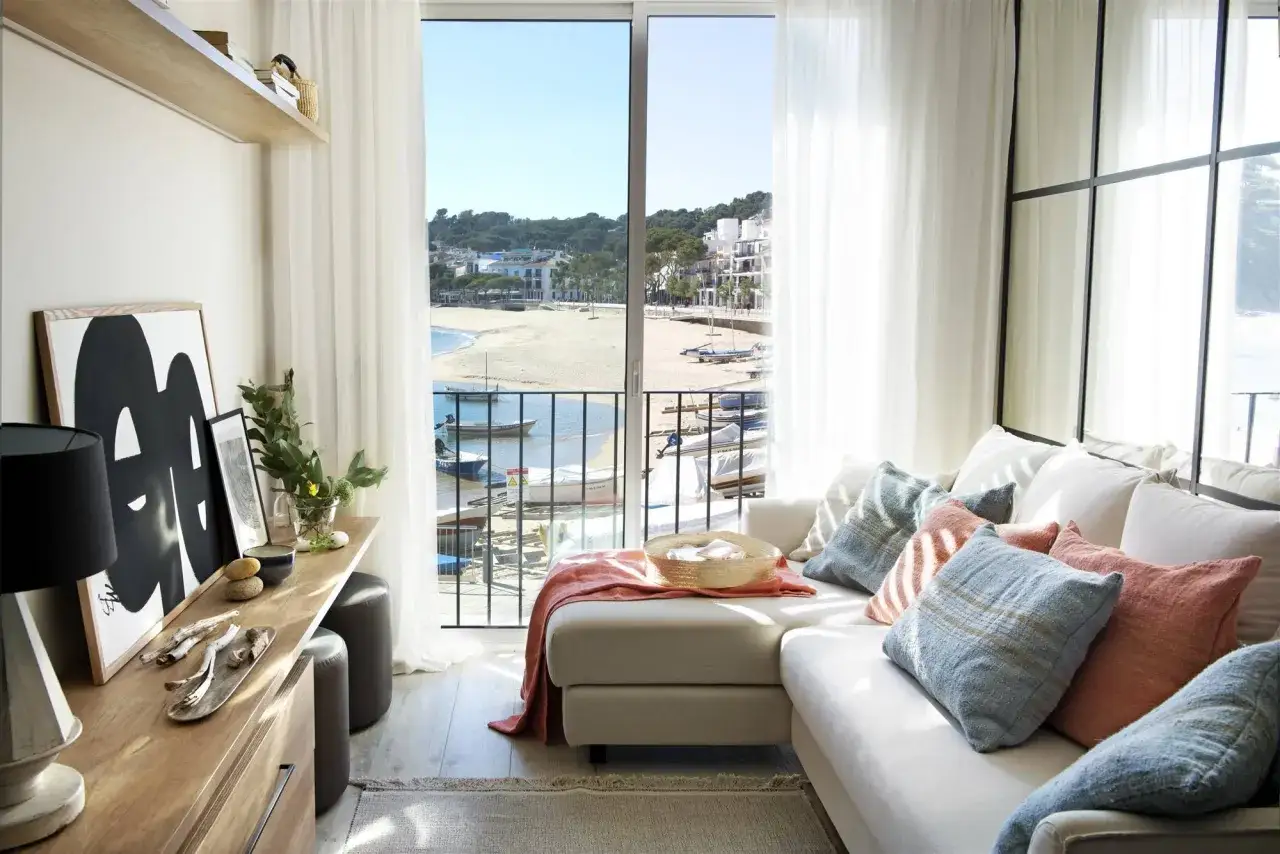 Salón estrecho y rectangular con sofá blanco, cojines de colores y vistas a una playa y un puerto con barcos.