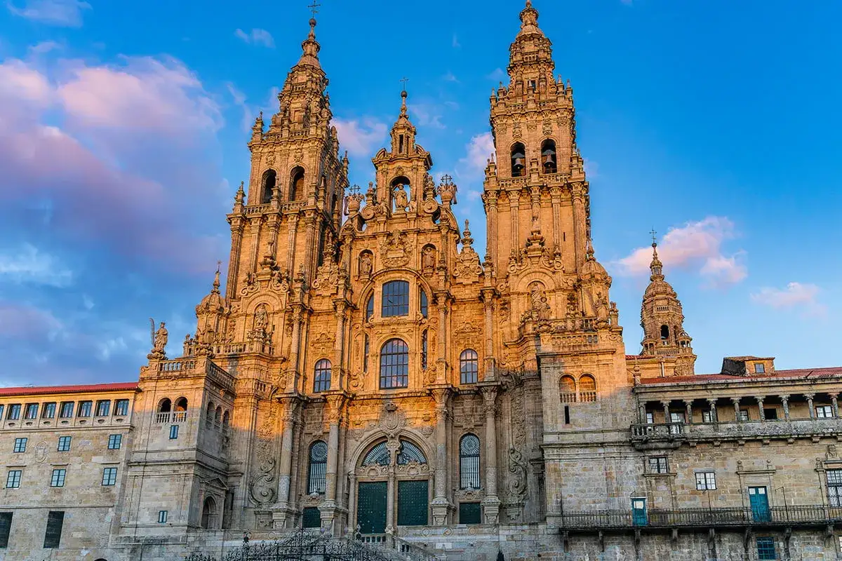 Majestatyczna katedra w Santiago de Compostela, oświetlona zachodzącym słońcem, z niebieskim niebem i chmurami w tle.