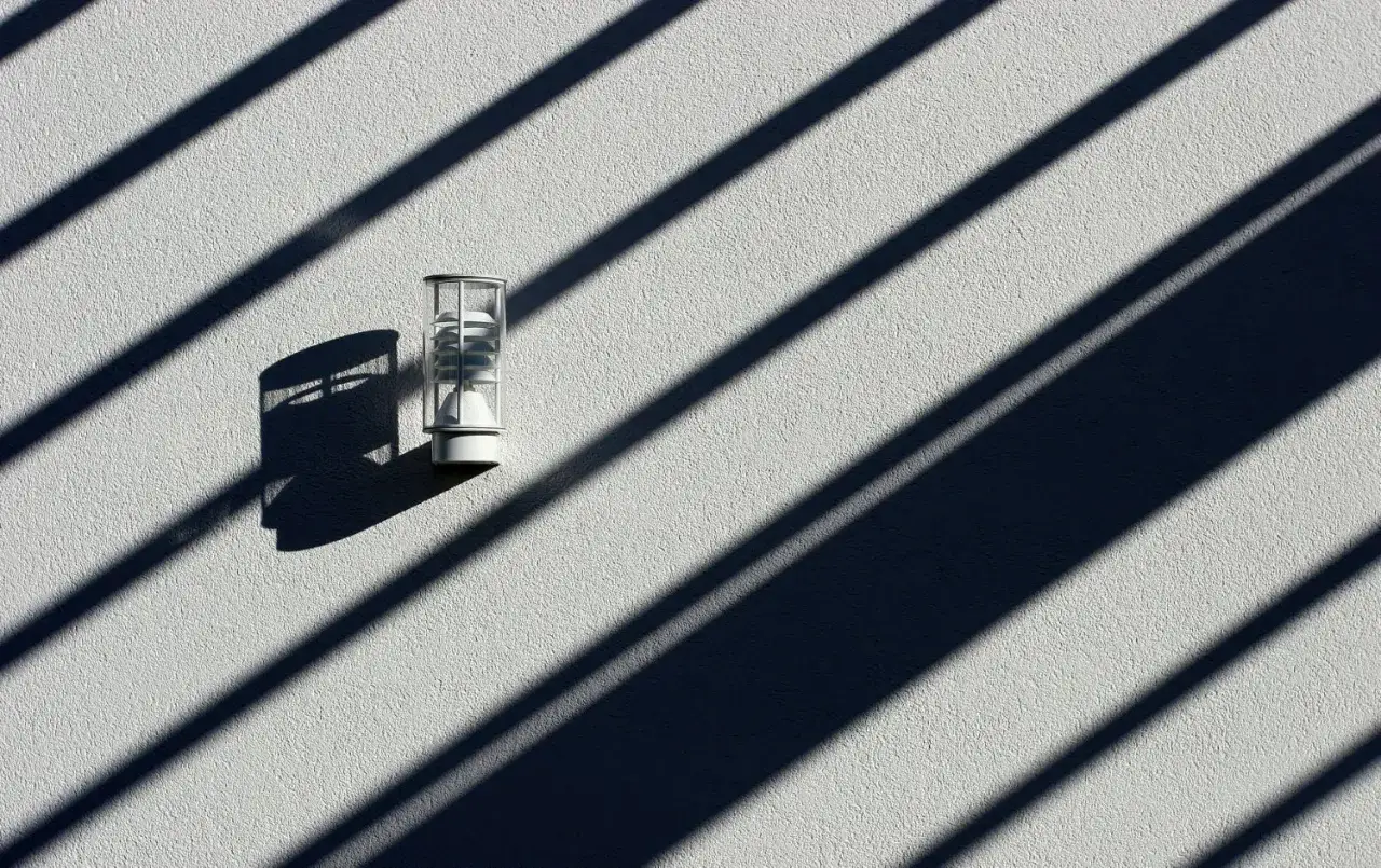 Een buitenlamp op een witte muur, met een dramatische **diagonaal compositie** van schaduwen van lamellen.