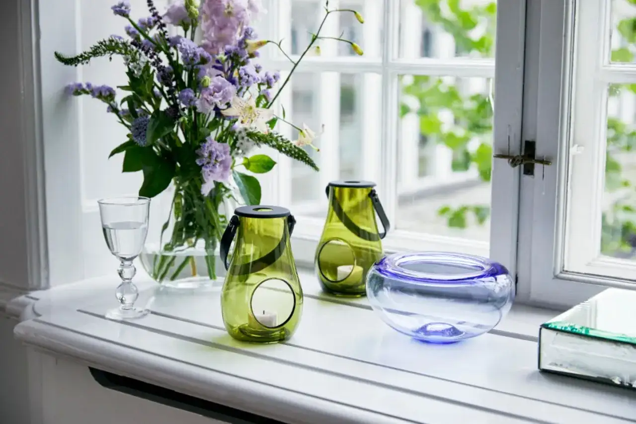 Ein Strauß lila Blumen, zwei grüne Windlichter und eine blaue Glasschale schmücken die Fensterbank. So lässt sich die Fensterbank dekorieren.