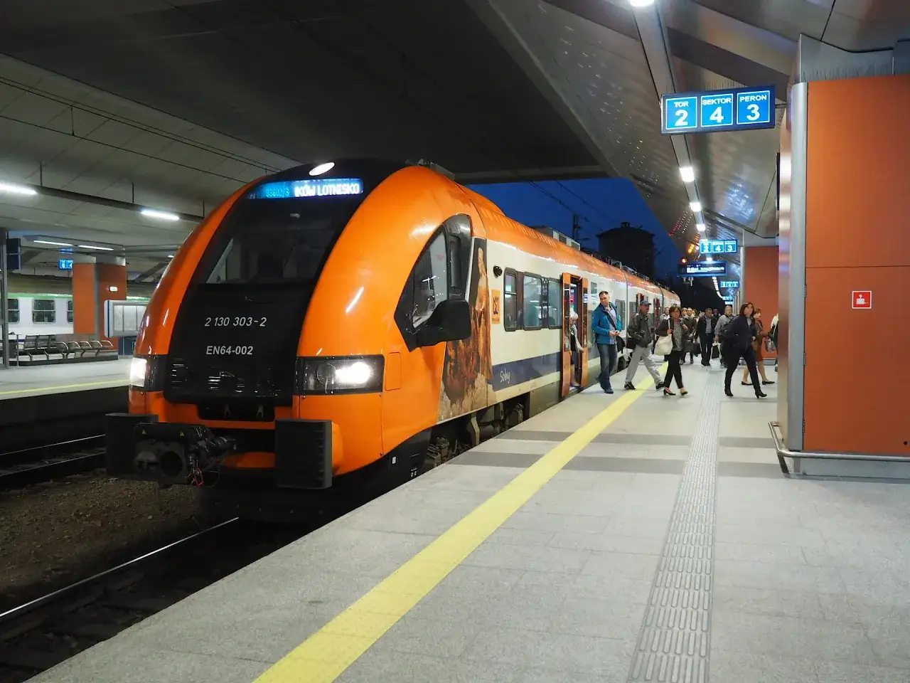 Pociąg PKP na stacji Kraków-Balice. Ludzie wsiadają i wysiadają, sprawdzając rozkład jazdy.