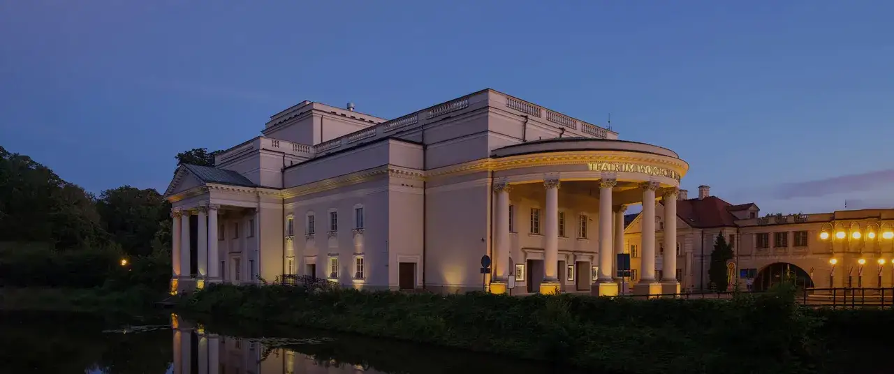 Elegancki budynek Teatru im. Wojciecha Bogusławskiego w Kaliszu, oświetlony wieczorem, odbija się w spokojnej wodzie.