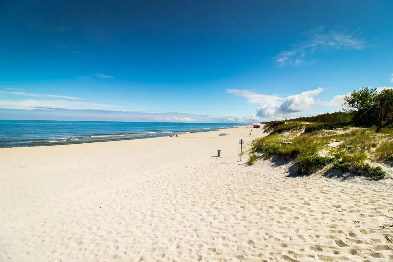 Plaża na Mierzei Wiślanej, z piaszczystymi wydmami i błękitnym niebem. W oddali widać kilka budek plażowych i drzewa.