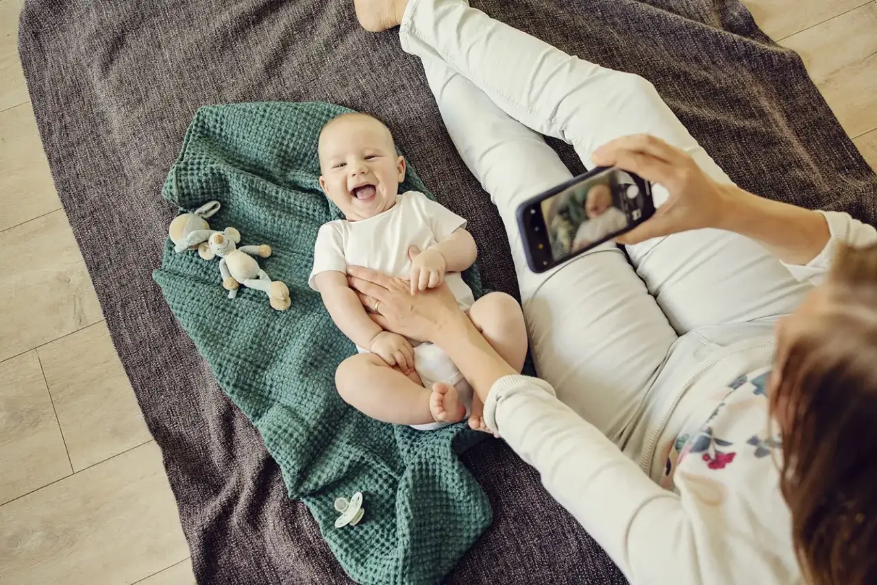 Uśmiechnięty noworodek przekręca się na bok na zielonym kocyku, a mama robi mu zdjęcie telefonem.