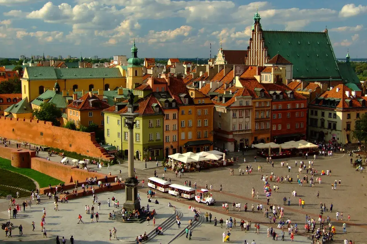 Tętniący życiem Rynek Starego Miasta w Warszawie. Kolorowe kamienice, Zamek Królewski i tłumy spacerowiczów tworzą malowniczy widok.